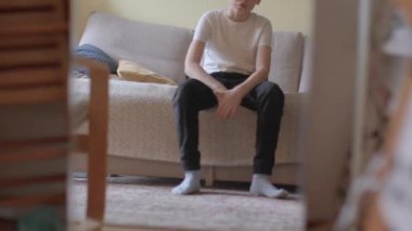 A young man dressed casually sits on a sofa in a warm and inviting living room, showcasing a relaxed and contemplative mood within a home setting.