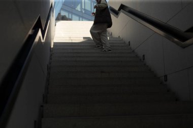 A person is carefully walking down a set of wide stairs, utilizing the handrail for support while casting a shadow on the steps below