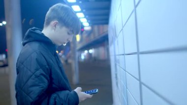 A young individual stands by a tiled wall during the nighttime, casually checking their phone and engaging with it