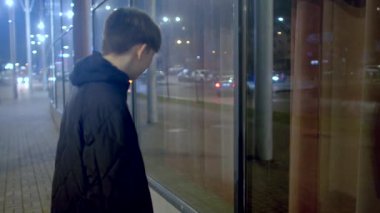 A young man stands alone at night, gazing out of a shop window, lost in thought. The moody atmosphere highlights themes of solitude