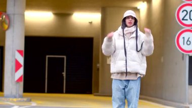 In a dim parking garage, two young women whisper while a man in a hood watches them and another man in a white jacket stands afar. Tension and intrigue build, suggesting an unfolding story