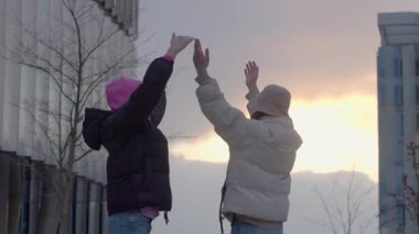 In this uplifting video, two young women joyfully stand together in front of a striking building, arms raised in celebration, capturing camaraderie