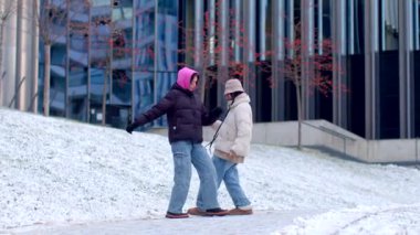 A man and woman enjoy a winter day, playing in the snow and walking in front of a picturesque building, filling the air with laughter and joy