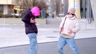 A lively street scene unfolds as two women in a pink hoodie stand on a bustling sidewalk, engaging with each other and a man who joins them