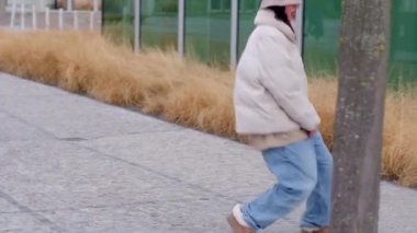A woman crouches by a tree on a city sidewalk, connecting with nature. A passerby strolls by, enhancing the tranquil atmosphere
