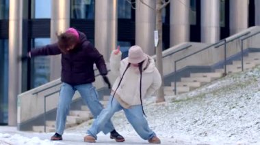 In this winter video, two women joyfully dance in the snow, creating a heartwarming scene by a charming building, filled with laughter and festive spirit