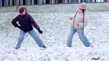 A joyful winter scene in a snowy park shows a couple strolling and laughing, two women having a snowball fight, and dancing in the snow