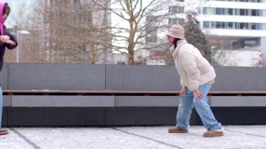 In a winter wonderland, two men dance in the snow, spreading joy. One reflects, another dances on a bench, while a couple shares warmth
