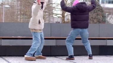 A busy sidewalk features interactions among people. Two men walk by a bench, a couple enjoys themselves, and a dancing couple attracts attention