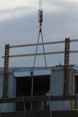İnşaat alanında ahşap kirişleri kaldıran bir vinç arka planda bulutlu bir gökyüzü olan bir binanın iskeletini sergiliyor..