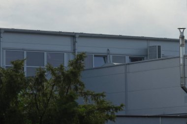 Çağdaş endüstriyel bir bina. Parlak bir tasarım, geniş pencereler ve bulutlu bir gökyüzünün altında yeşilliklerle çevrili kendine özgü bir çatı çizgisi var..