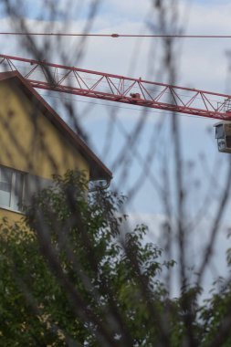 A red construction crane towers over a residential building, framed by trees, showcasing urban development against a cloudy sky.