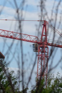 A striking red construction crane stands tall against a backdrop of clouds, framed by branches, symbolizing ongoing development in an urban environment.
