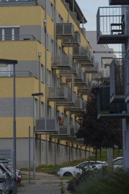 A contemporary multi-story apartment complex showcasing balconies and nearby parked cars, surrounded by greenery and urban scenery.