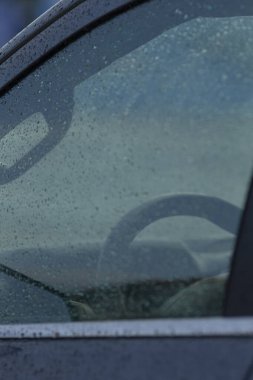 Yağmur damlalarının ve direksiyonun yansımasının yer aldığı bir arabanın yakın çekim görüntüsü, sakin ve atmosferik bir manzara yaratıyor..