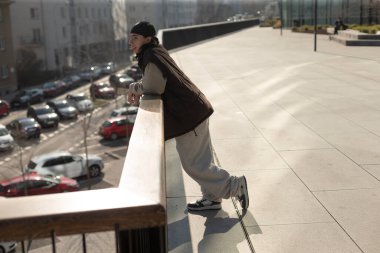 A man is casually leaning against a railing while looking out over a large parking lot filled with parked cars and shadows
