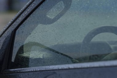 A close-up view of a car interior with a rain-soaked window, capturing the reflections of the steering wheel and side mirror. A moody and atmospheric shot.