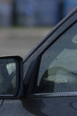Pencerede yağmur damlalarını gösteren siyah bir arabanın yakın çekimi. Görüntü kasvetli bir atmosferi yansıtıyor ve şehir yaşamını yansıtıyor..