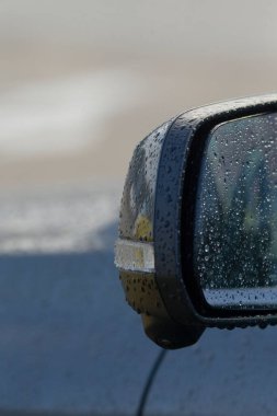 Yağmurlu çevreyi yansıtan bir araba yan aynası, yüzeyde yağmur damlaları ve bulanık bir arka plan görüntüsü..