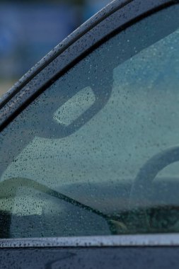 Yağmurdan sırılsıklam olmuş bir arabanın yakın plan çekimi camdaki damlacıkları ve soyut yansımaları sergiliyor ve kasvetli bir atmosfer yaratıyor..