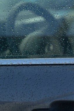 Yağmurdan sırılsıklam olmuş bir arabanın penceresinin yakın çekimi, içerideki direksiyonun bulanık bir yansımasını ortaya çıkarıyor, dengesiz ve atmosferik bir etki yaratıyor..