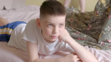 A young boy in casual attire looks pensive while lying on a bed, displaying various emotions. This captures the essence of childhood contemplation.