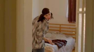 A woman in a plaid shirt stands in a cozy, warmly lit bedroom beside a neatly made bed. The scene radiates tranquility and relaxation
