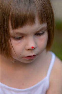There is a cute little girl who has a charming red heart painted on the bridge of her nose, adding to her delightful appearance