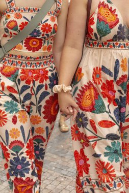 Two women dressed in vibrant, floralpatterned dresses are joyfully holding hands as they stroll down the busy street together