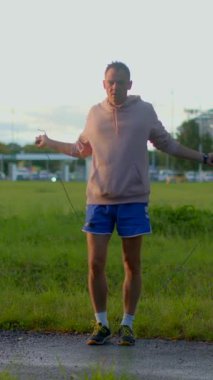 A man is seen joyfully skipping rope in a lush grass field, dressed in casual shorts and a comfortable hooded sweatshirt, engaging in his fitness routine outdoors on a bright, sunny day
