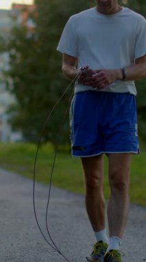 This video shows a man in a white shirt and blue shorts doing an outdoor workout. He begins with stretches, then walks with a jump rope, emphasizing his commitment to fitness