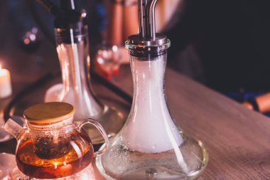 Masada mumlar ve dumanla Nargile manzarası arka planda sigara içen gençlerle nargile salonu, Shisha parti kulübü konsepti.