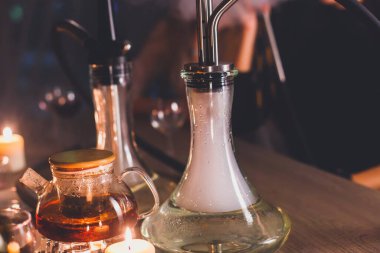 Masada mumlar ve dumanla Nargile manzarası arka planda sigara içen gençlerle nargile salonu, Shisha parti kulübü konsepti.