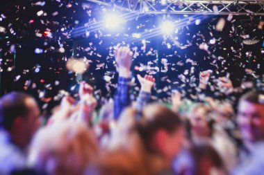 Bir konser festivali sırasında dans pistinde renkli konfeti patlaması, sahne ışıklarıyla dolu kalabalık konser salonu, rock gösterisi performansı, insanların silueti.
