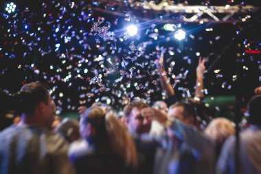 Bir konser festivali sırasında dans pistinde renkli konfeti patlaması, sahne ışıklarıyla dolu kalabalık konser salonu, rock gösterisi performansı, insanların silueti.