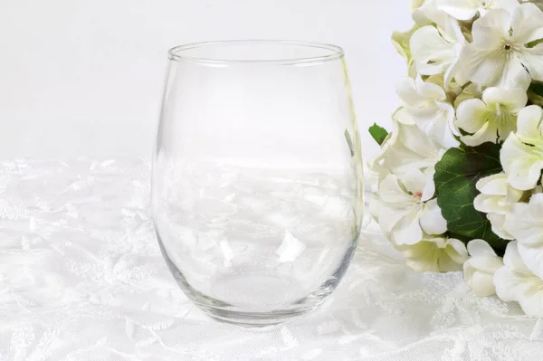 Stemless wine glass atop a white lace background surrounded by cheery white hydrangeas.