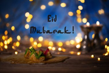 Kutlama: EID mübarek! Ahşap bir yüzey üzerinde Arapça tatlılar. Mumluk, gece lambası ve gece mavi gökyüzü ile arka planda hilal ay.