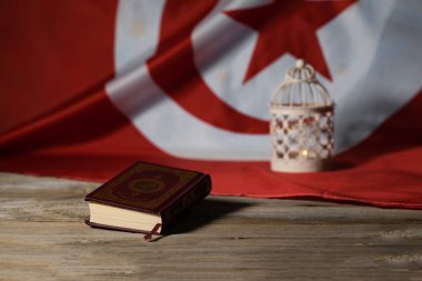 Sacred book of Quran on a wooden surface. Tunisian flag and candle holder in the background. Translation - the book contains verses of Koran clipart