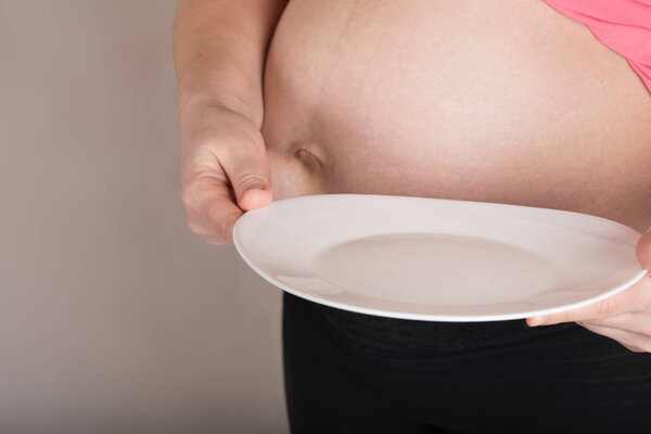 Young pregnant woman keeps empty white plate close to her belly.