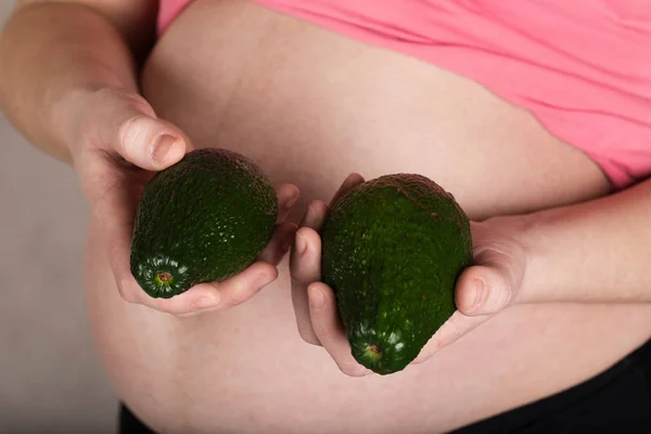 Молодая беременная женщина держит два авокадо близко к животу . — стоковое фото
