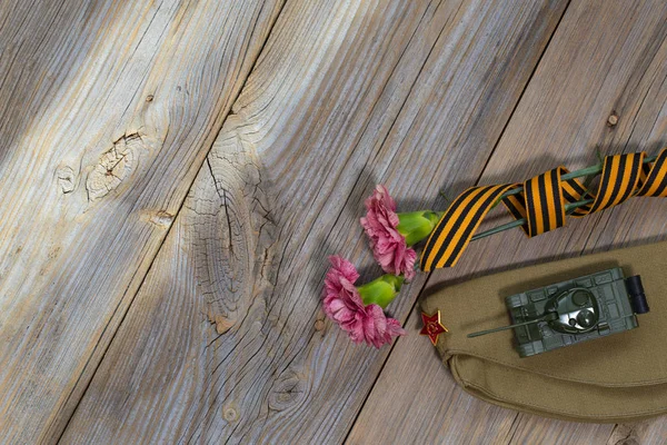 Zwei rosa Nelken, Saint George Band, Tank und Militärische Kappe auf einer Holzoberfläche. — Stockfoto