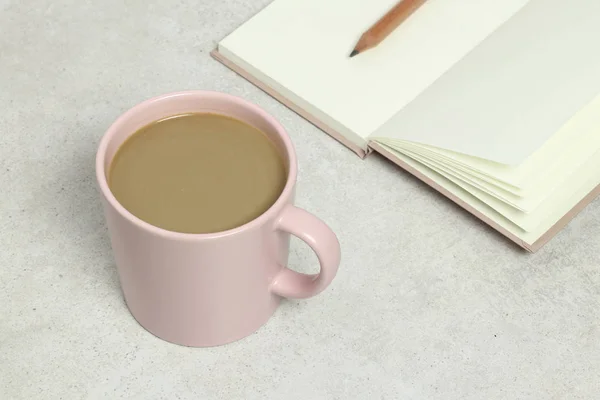 The pink cup of coffee, opened book and pencil on the grey granite texture