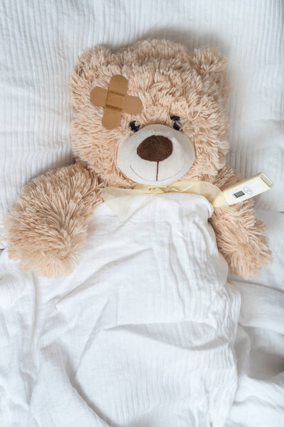 Sick Teddy bear with plaster and thermometer is lying in bed.