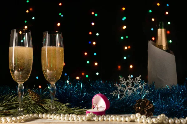 Christmas and New Year dinner of a couple with two glasses of champagne and a wedding ring. Marriage proposal concept. Pearls, tinsel and spruce branch on a black background with blurred lights.