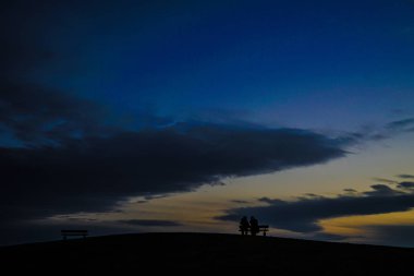 Ebeveynler ve çocuklar bankın tepelerinde gün batımında oturacaklar.