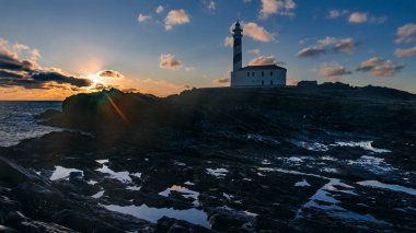 İspanya, Menorca 'da bulutlu gökyüzü ile kış günü şafak vaktinde güneş patlamaları olan deniz feneri