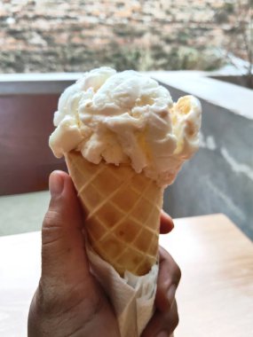 Bir el, ılık ahşap bir zemine karşı lezzetli beyaz bir dondurma külahı tutar, ferahlatıcı bir yaz ziyafeti için mükemmeldir..