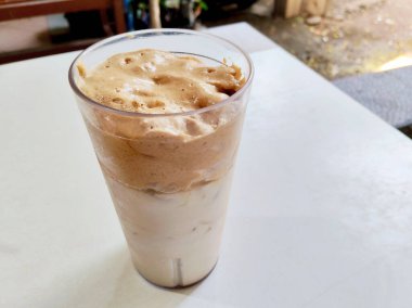 Bir bardak ev yapımı Dalgona kahvesi, kremalı popüler bir kahve, beyaz bir masada buzlu sütün üzerine köpüklü bir köpük..