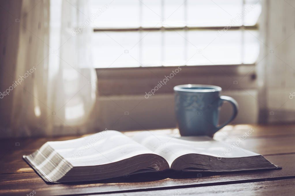 Close up of open bible with a cup of coffee on wooden table against window light