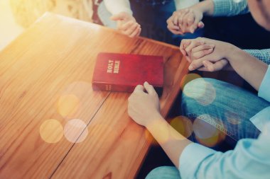 Close up of  people group holding a hand and pray together over a blurred holy bible on wooden table, Christian fellowship  or praying meeting in home concept with copy space clipart